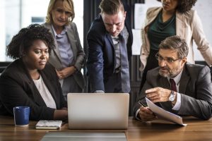 A diverse team of four professionals in business attire gather around a desk to review documents and a laptop