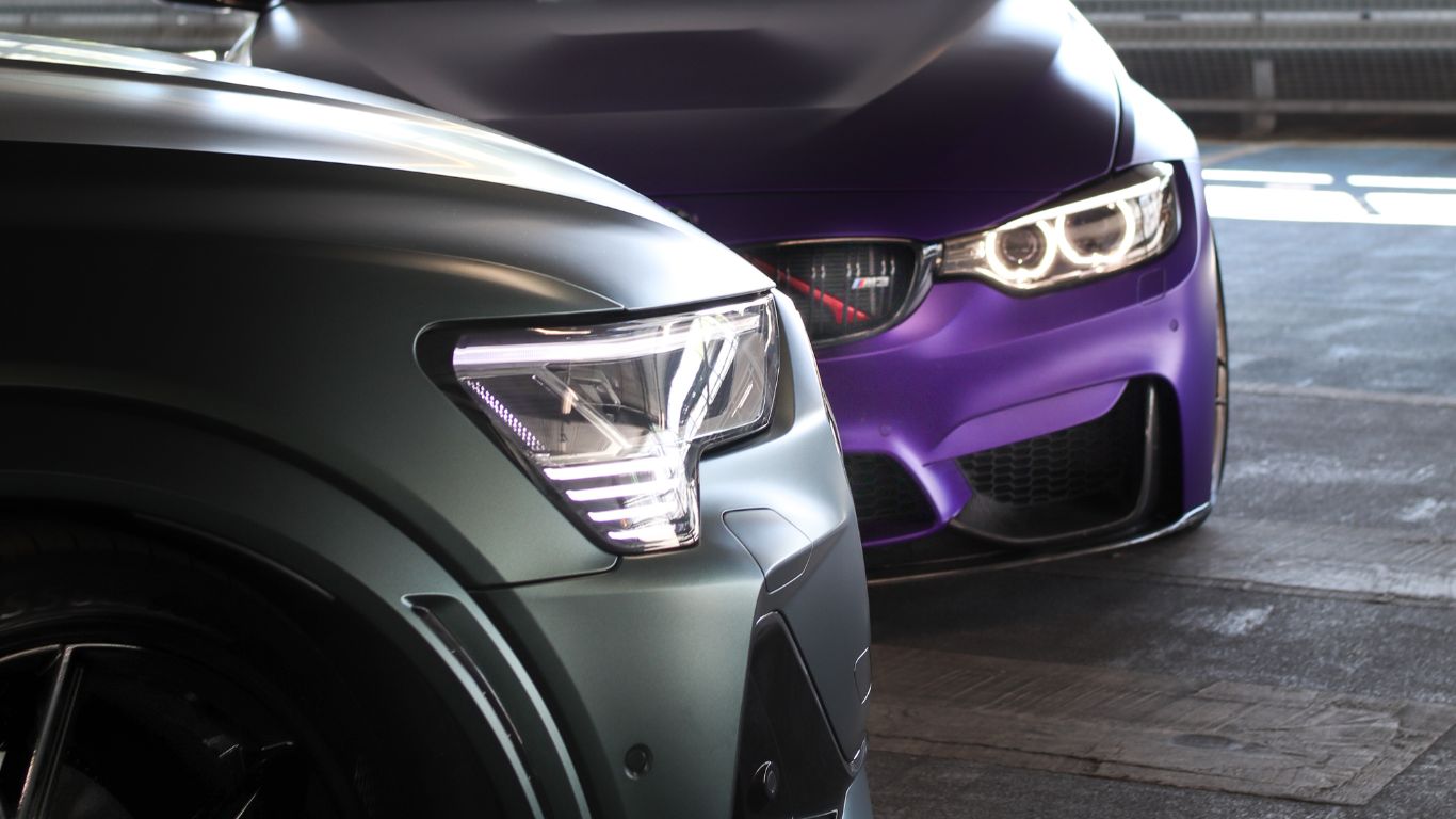 Close-up of the front headlights and hoods of two modern cars, one grey and one purple, parked side by side