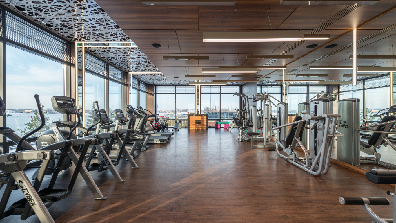 Modern gym interior with rows of cardio machines and weights facing large windows overlooking a waterfront