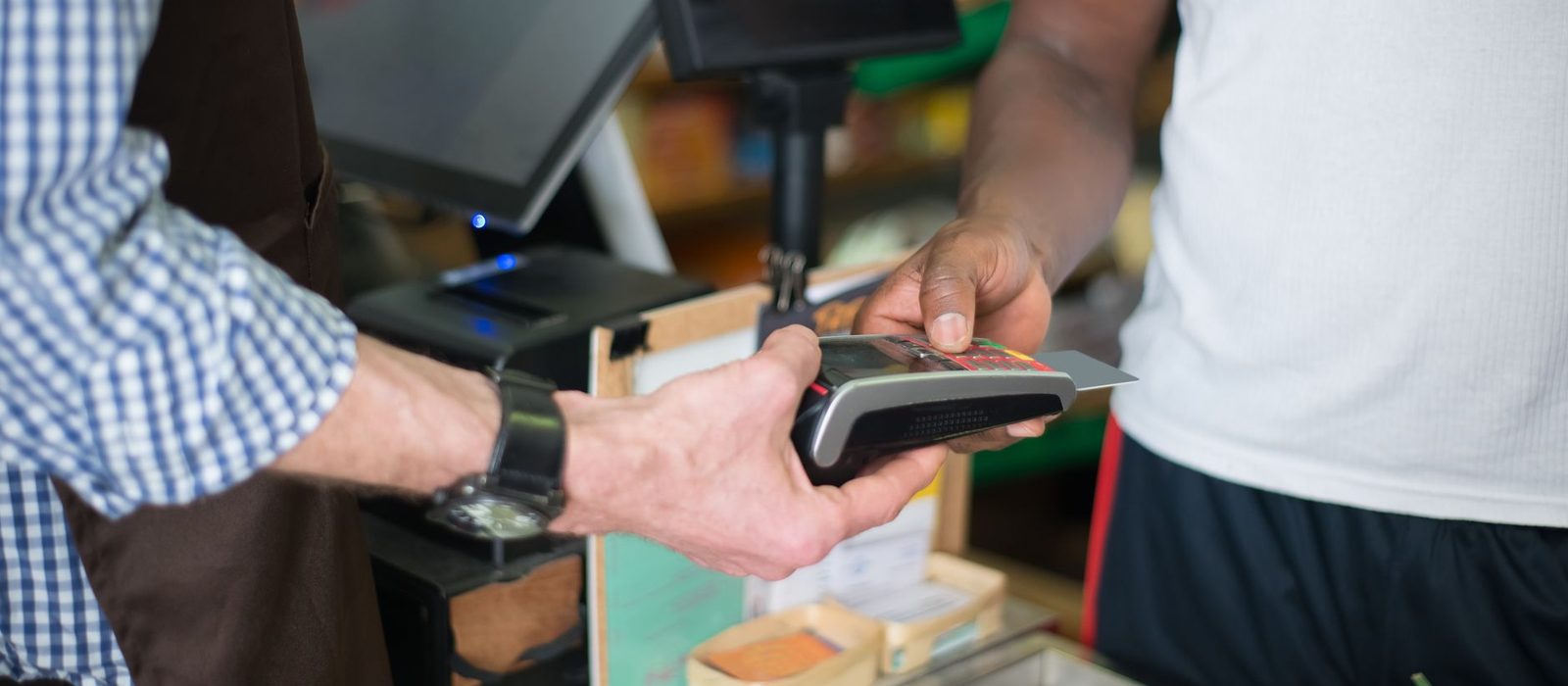 A customer taps a bank card on a handheld terminal held by a grocer at the counter
