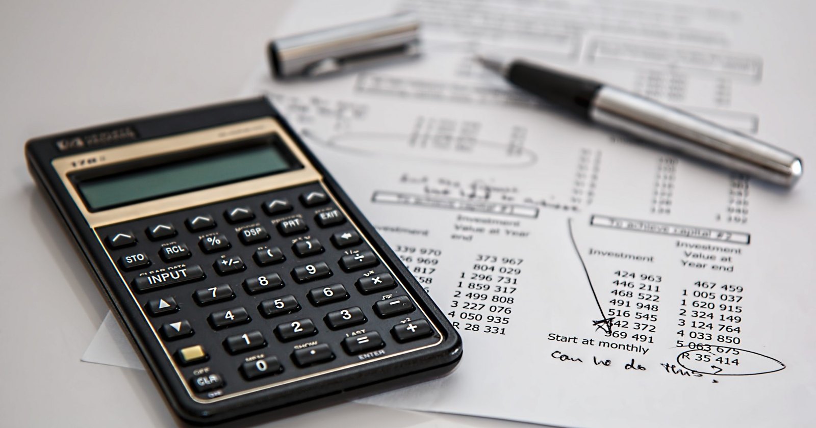 A calculator resting on printed investment projections, with a pen and a handwritten note reading can we do this