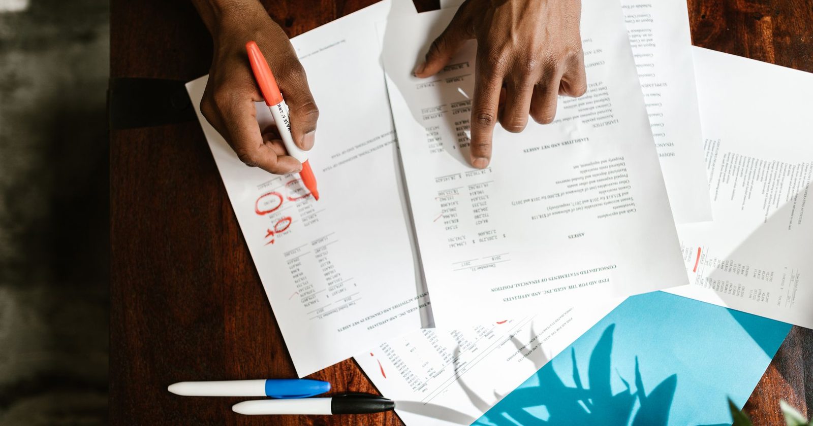 Two people lean over printed financial statements on a desk circling figures with a red marker