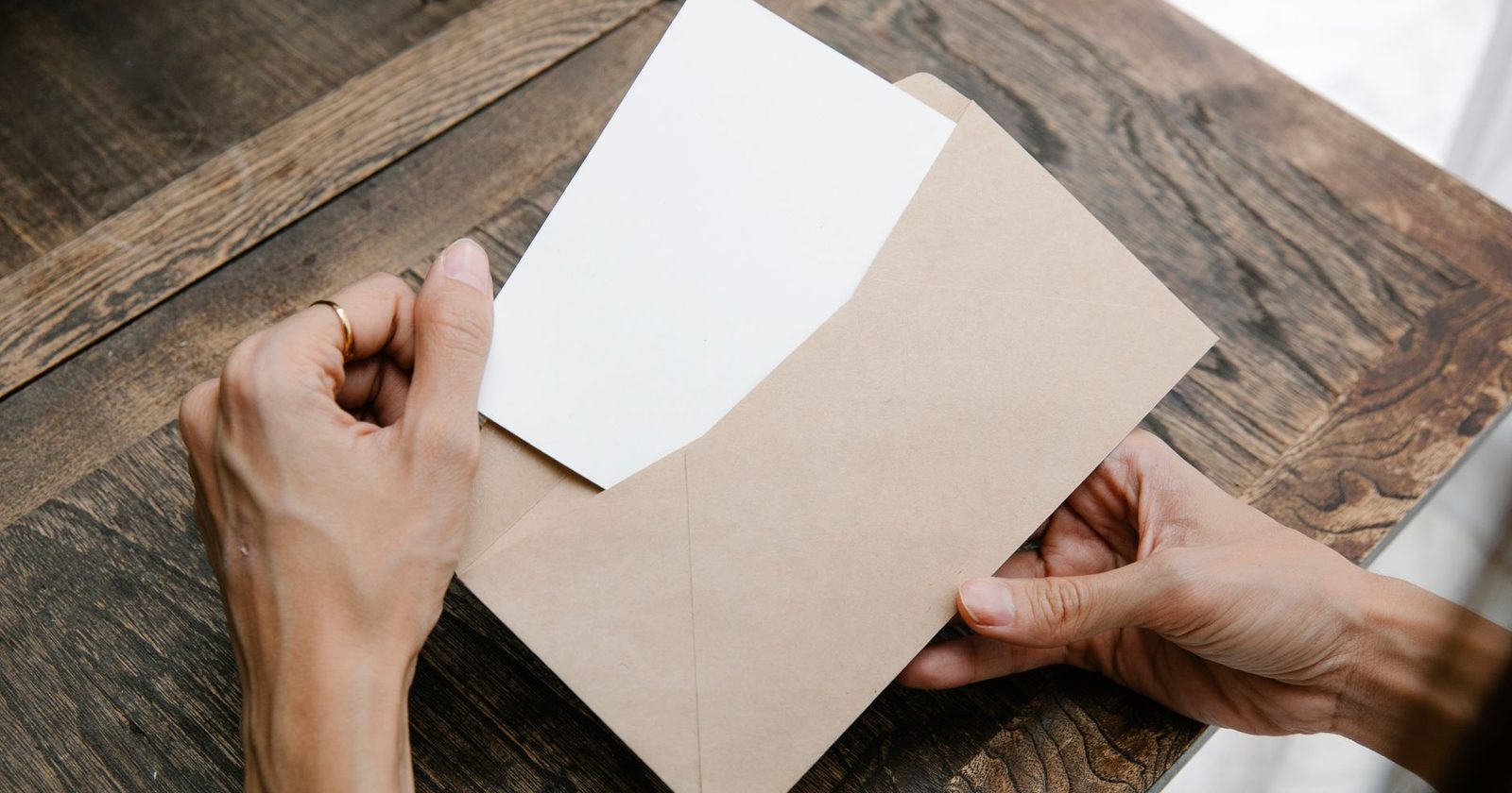 A pair of hands sliding a letter out of a kraft paper envelope on a dark timber table.