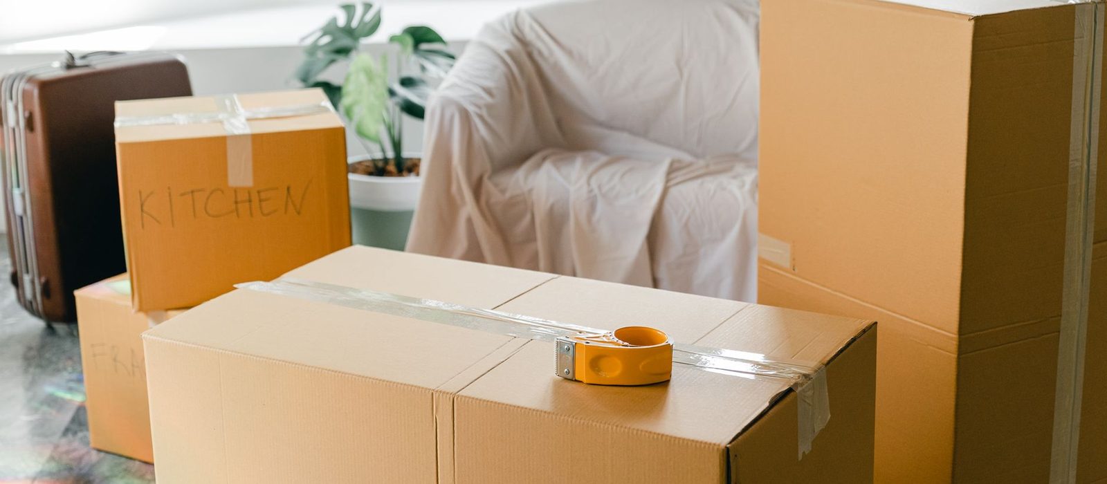 Taped cardboard moving boxes stacked in a bare room, one marked Kitchen, beside an armchair under a dust sheet.