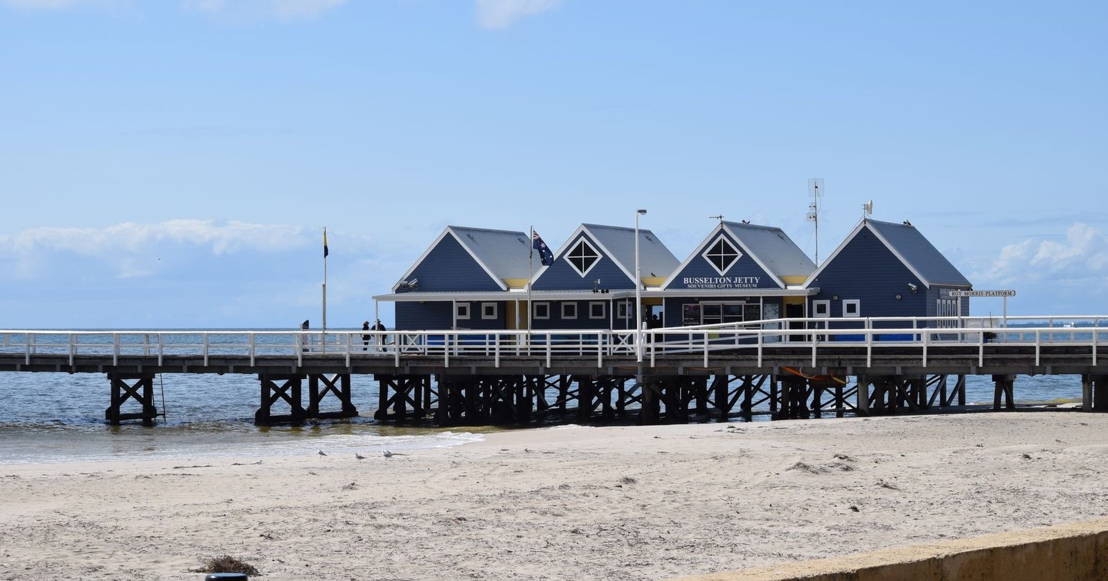 Blue timber sheds on the Busselton Jetty with a white railing, seen from the beach on a clear day